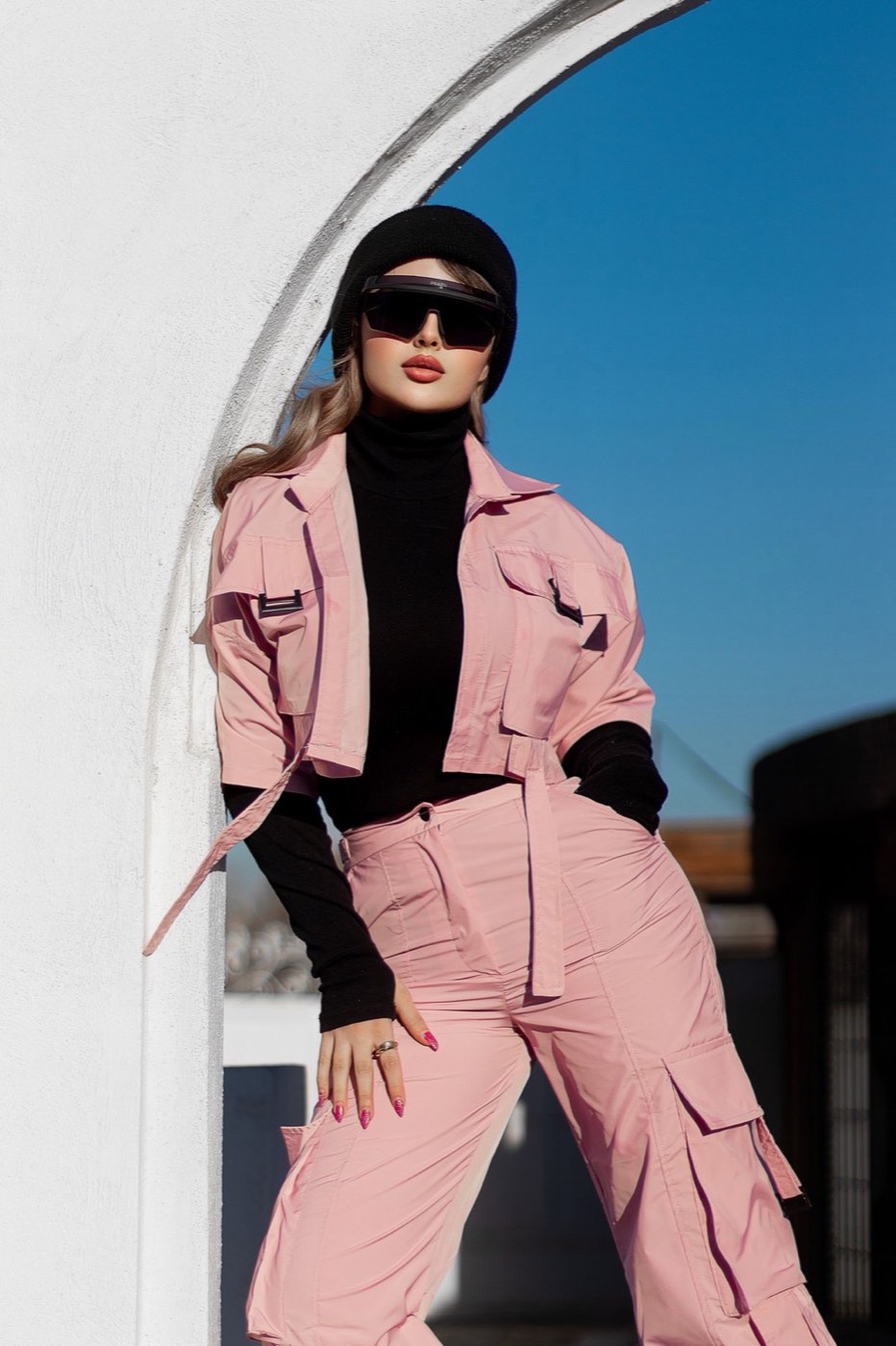 A person in a pink outfit standing against a white wall in bright daylight.