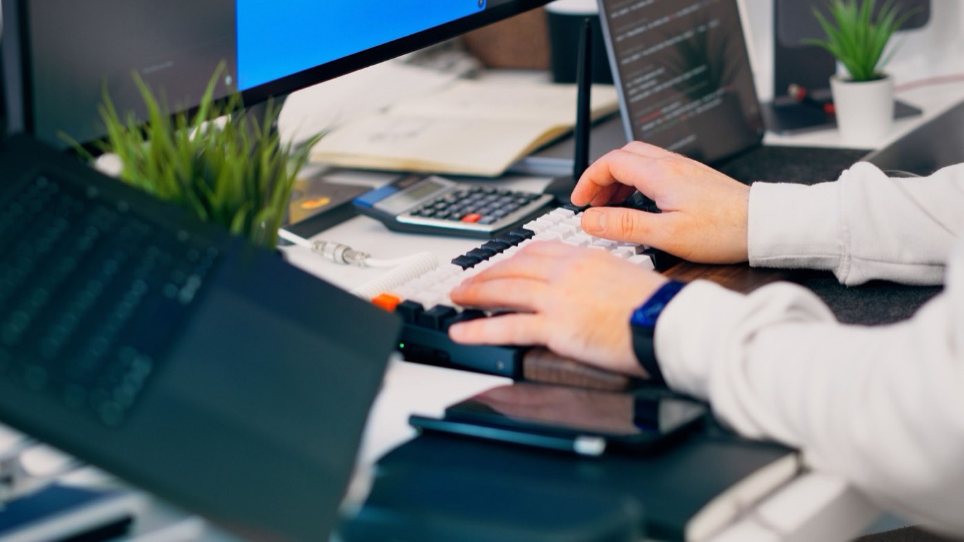 Hands typing at a desk with two monitors and a code editor open.