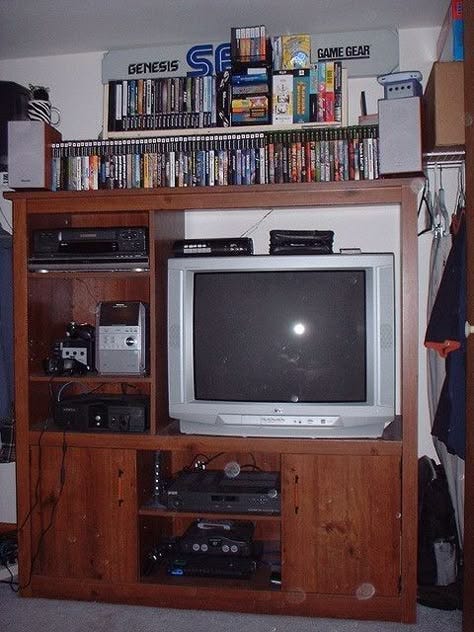 A retro home gaming setup: a CRT TV in a wooden cabinet beneath shelves lined with Sega Genesis, Game Gear, and other console games.
